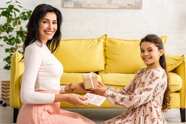Cute, elegant child presenting mothers day card and gift box to happy mom while sitting on floor together — Stock Photo