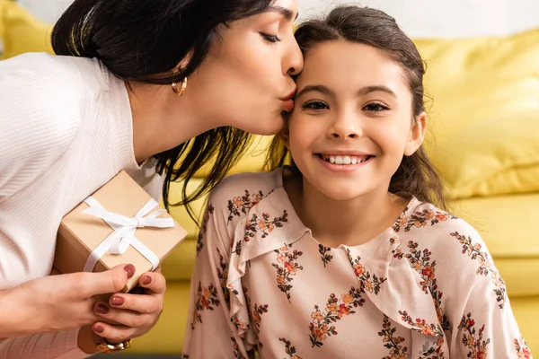 Allegra figlia sorridente alla macchina fotografica mentre mamma felice che tiene la scatola regalo e la bacia — Foto stock