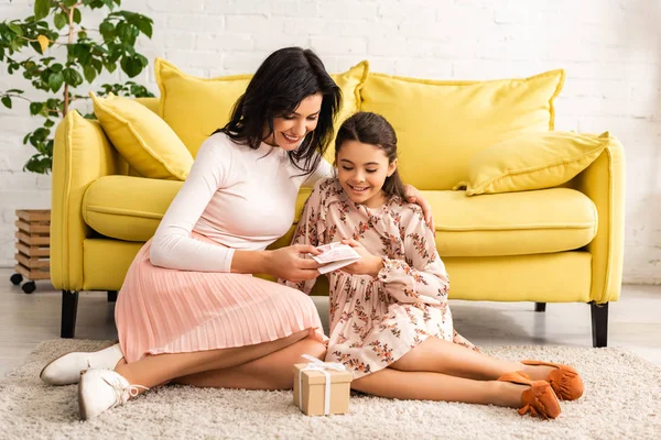 Fille élégante présentant la carte de jour des mères à maman heureuse tout en étant assis sur le sol près du canapé jaune ensemble — Photo de stock
