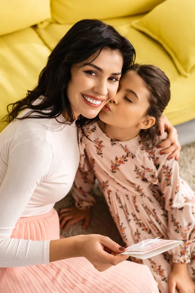 Mujer feliz mirando a la cámara mientras sostiene la tarjeta del día de las madres mientras que la hija adorable besándola - foto de stock