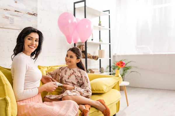 Adorable hija presentando las madres tarjeta de día a la mamá feliz mientras se sienta en el sofá amarillo juntos - foto de stock