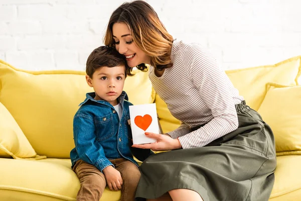 Donna allegra che abbraccia adorabile figlio mentre tiene il biglietto di giorno di madri con simbolo di cuore — Foto stock