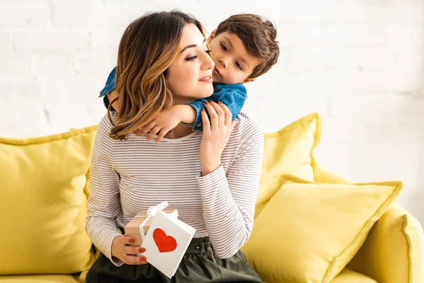 Niedlicher Junge umarmt glückliche Mutter mit Geschenkbox und Muttertagskarte mit Herzsymbol — Stockfoto