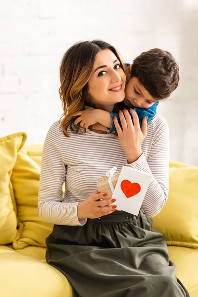 Donna felice guardando la macchina fotografica mentre tiene in mano la scatola regalo e la carta di giorno delle madri con il simbolo del cuore, mentre adorabile figlio la abbraccia — Foto stock