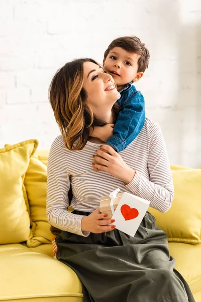 Lindo niño abrazando feliz madre sosteniendo caja de regalo y la tarjeta de día de las madres con símbolo del corazón - foto de stock