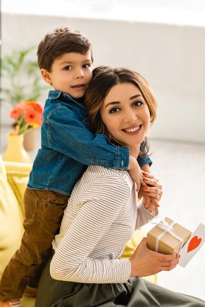 Cute boy embracing mom holding gift box and mothers day card with heart symbol — Stock Photo