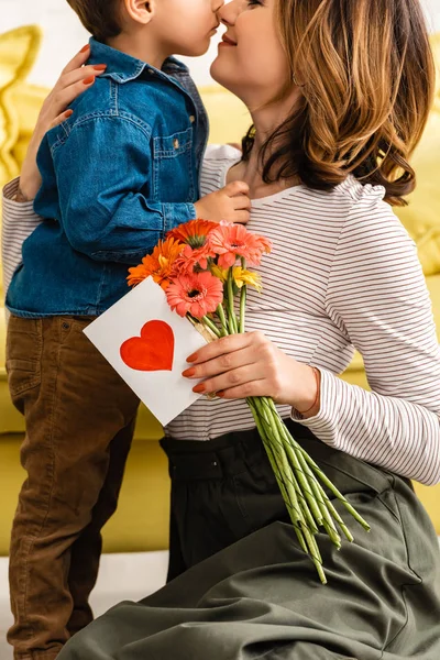 Cropped view of happy woman holding flowers and mothers day card with heart symbol while embracing adorable son — Stock Photo