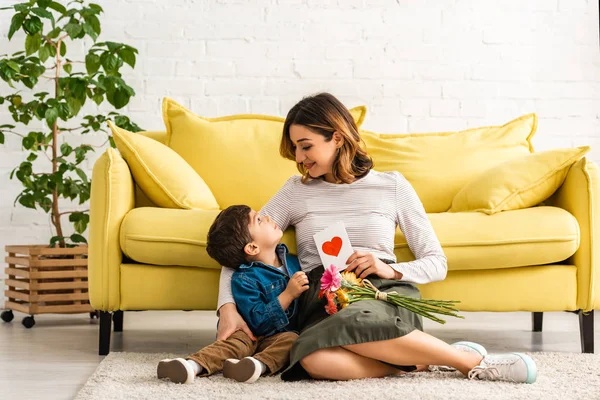 Smiling woman looking at adorable son while sitting on floor with mothers day card and flowers — Stock Photo