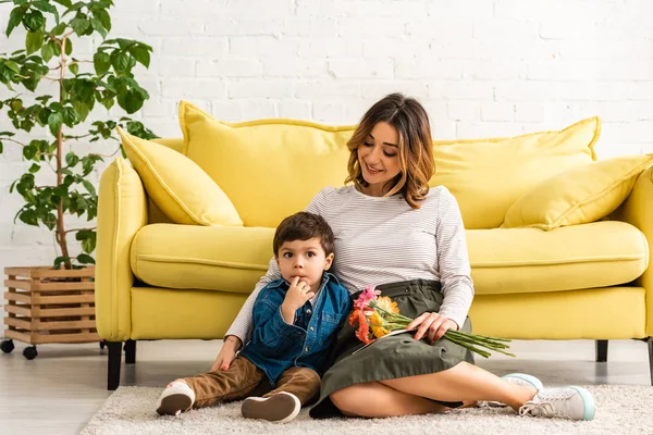 Glückliche Frau, die am Muttertag mit Blumen auf dem Boden sitzt und ihren entzückenden Sohn umarmt — Stockfoto