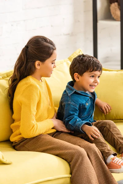 Alegre chico sentado en amarillo sofá cerca adorable hermana en casa - foto de stock