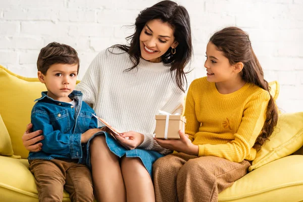 Mujer alegre sosteniendo la tarjeta del día de las madres mientras que abraza al hijo, mientras que la hija sonriente sostiene la caja de regalo - foto de stock