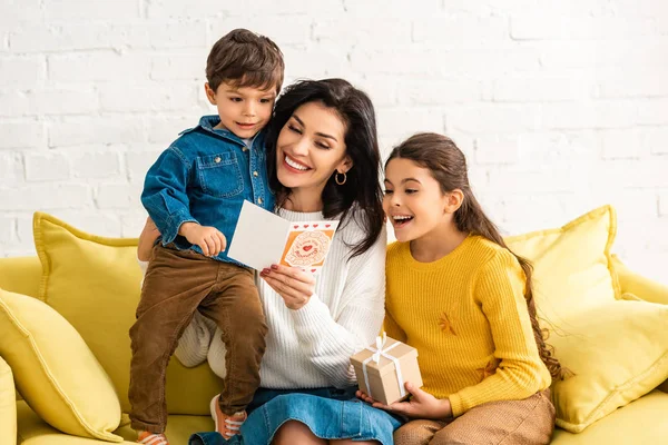 Femme heureuse tenant carte de jour de mères et embrassant fils, tandis que la fille tenant boîte cadeau — Photo de stock