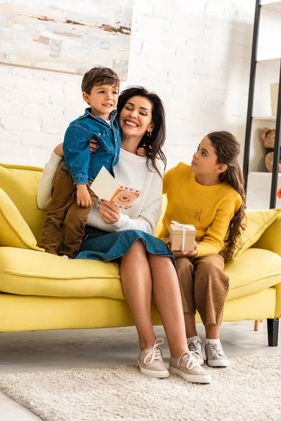 Mujer feliz sosteniendo la tarjeta del día de las madres y abrazando al hijo, mientras que la hija sostiene la caja de regalo - foto de stock