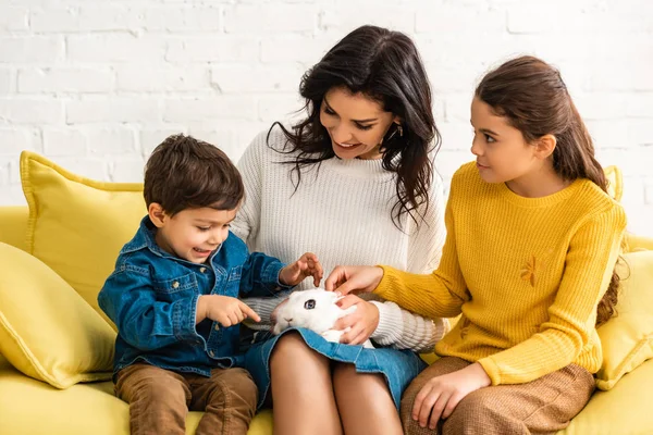 Fröhliche Mutter und Kinder auf gelbem Sofa mit weißem Kaninchen — Stockfoto