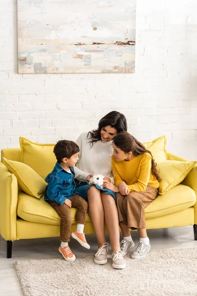 Mère heureuse et les enfants assis sur un canapé jaune avec lapin blanc — Photo de stock