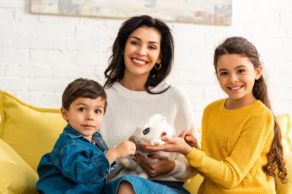 Happy children and cheerful mother looking at camera while sitting on sofa with white funny rabbit — Stock Photo
