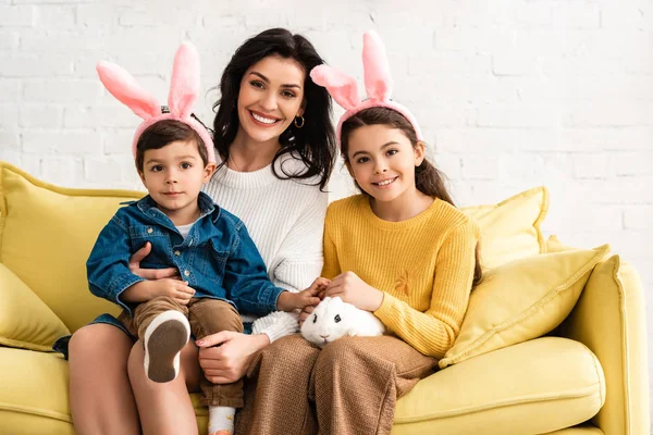 Счастливая женщина и веселые дети в кроликовых ушах улыбаются в камеру, сидя на желтом диване с белым смешным кроликом — стоковое фото