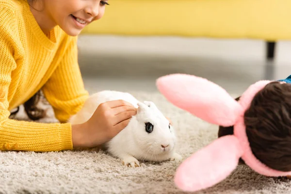 Vue recadrée des enfants couchés sur le sol près du lapin drôle blanc — Photo de stock