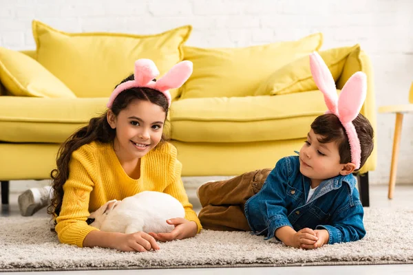 Adorable kids in bunny ears lying on floor near white funny rabbit — Stock Photo