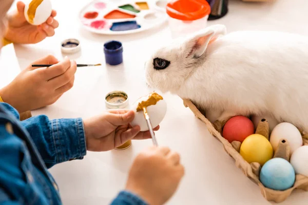 Cropped view of kids painting Easter eggs while sitting at desk near white rabbit — Stock Photo
