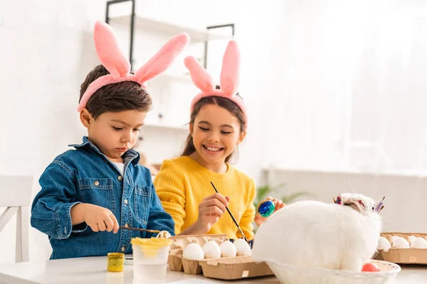 Enfants gais dans des oreilles de lapin peignant des œufs de Pâques assis au bureau près du lapin blanc en osier — Photo de stock