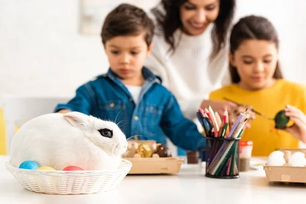 Enfoque selectivo de conejo blanco en mimbre cerca de la madre y los niños pintando huevos de Pascua - foto de stock