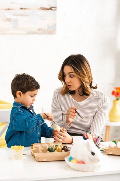 Внимательная мать и сын рисуют пасхальные яйца, сидя за столом рядом с кроликом в плетеном — стоковое фото