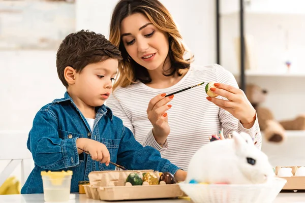 Happy mother and son painting Easter eggs while sitting at table near bunny in wicker — Stock Photo