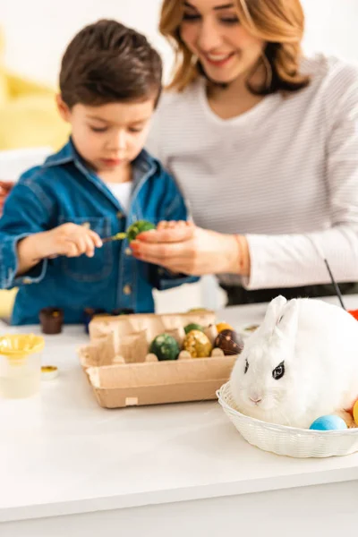 Messa a fuoco selettiva di madre e figlio pittura uova di Pasqua mentre seduto a tavola vicino coniglietto in vimini — Foto stock