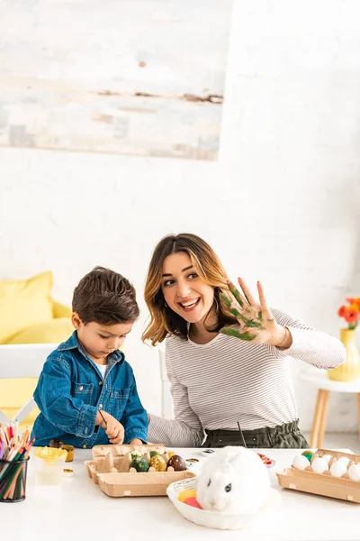 Femme joyeuse agitant main peinte tout en peignant des œufs de Pâques avec son fils — Photo de stock