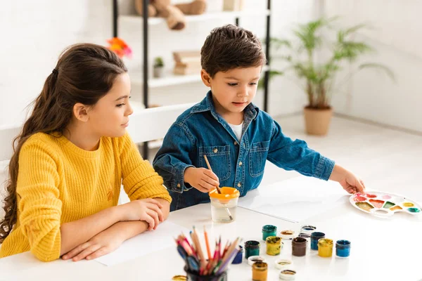 Cute boy dipping paintbrush in water and touching palette while sitting at table near sister — Stock Photo