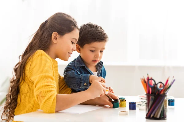 Fratello e sorella concentrati seduti a tavola e a disegnare con pitture insieme — Foto stock