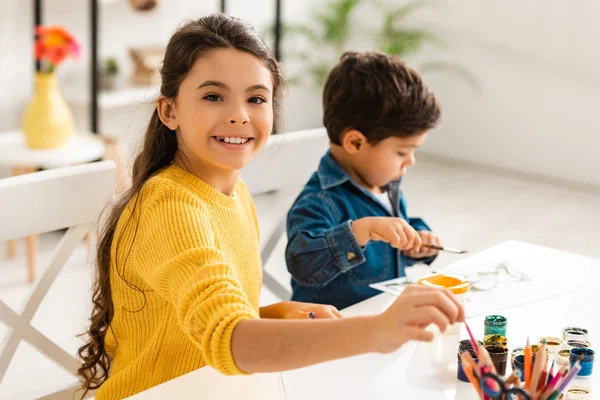 Fröhliche Schwester blickt in die Kamera, während sie am Tisch sitzt und mit ihrem Bruder zeichnet — Stockfoto