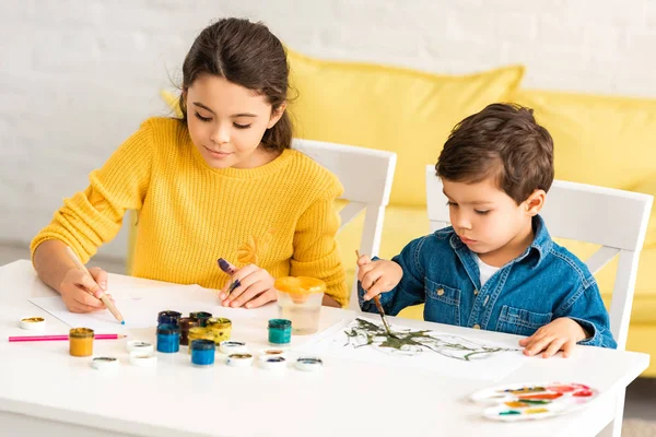 Crianças atenciosas e adoráveis sentadas à mesa e desenhando com tintas juntas — Fotografia de Stock