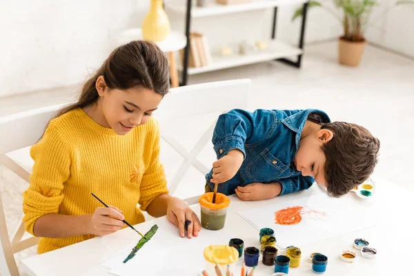 Menino bonito misturando tintas em recipiente enquanto sentado perto irmã desenho com pincel — Fotografia de Stock