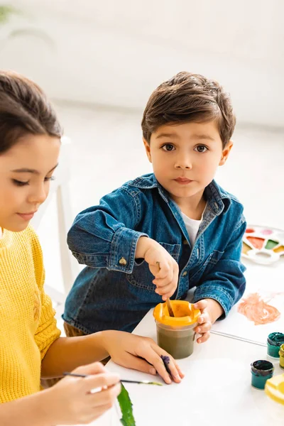 Adorable garçon mélange des peintures dans le récipient tout en étant assis près de sœur et en regardant la caméra — Photo de stock