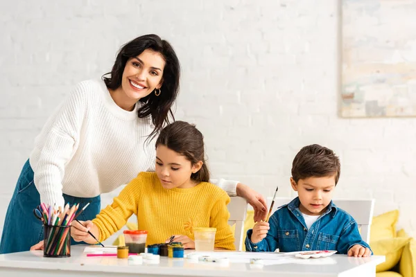 Glückliche Frau, die am Schreibtisch steht und in die Kamera lächelt, während entzückende Kinder mit Farben zeichnen — Stockfoto