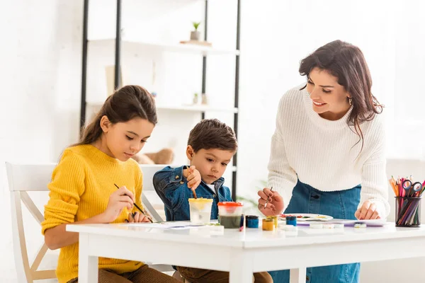 Щаслива жінка стоїть біля столу і малює з фарбами разом з дітьми — стокове фото