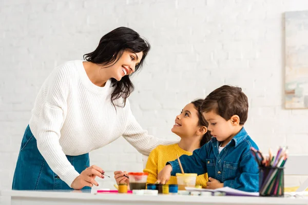 Happy mother looking at children sitting at table and drawing with paints — Stock Photo