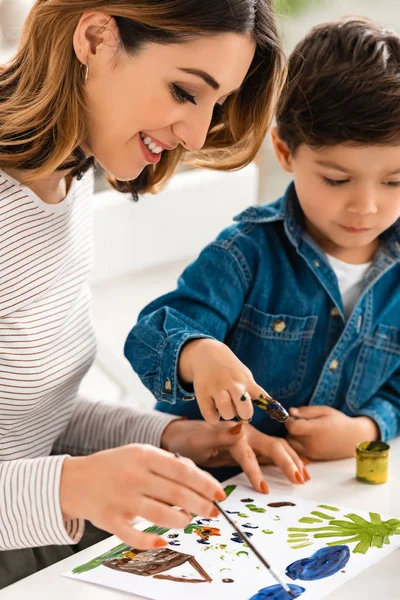 Mujer feliz sentada en la mesa y dibujando con pinturas junto con el hijo - foto de stock