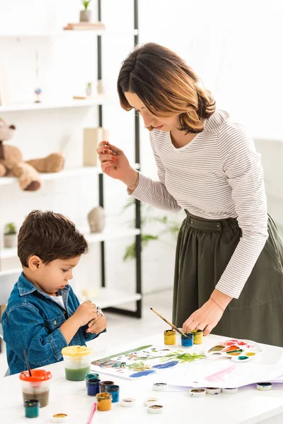 Junge Mutter steht am Tisch, während süßer Sohn Container mit Farben hält — Stockfoto