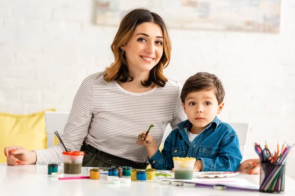 Feliz madre e hijo mirando a la cámara mientras dibujan con pinturas juntos - foto de stock