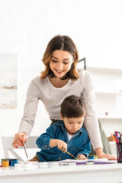 Happy mother holding paintbrush while son drawing with paints — Stock Photo