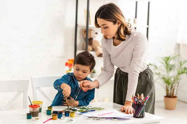 Jovem mãe segurando pincel enquanto filho desenho com tintas — Fotografia de Stock