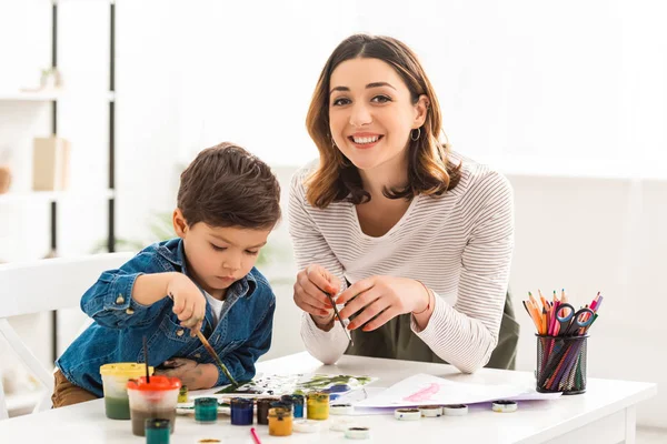 Mujer feliz mirando a la cámara mientras dibuja con pinturas junto con el hijo - foto de stock