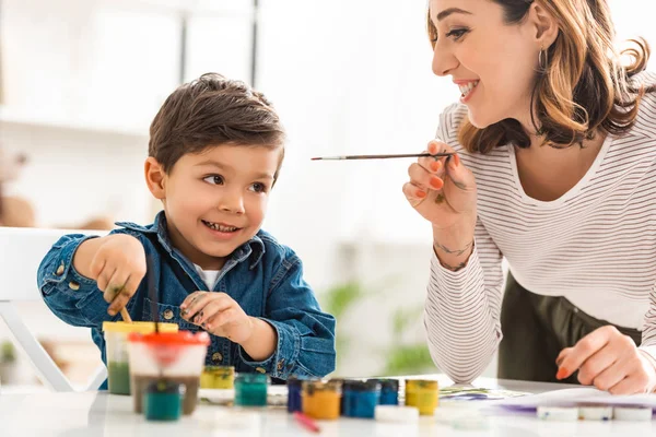 Joyeuse mère et son fils tenant pinceaux près des conteneurs avec des peintures — Photo de stock