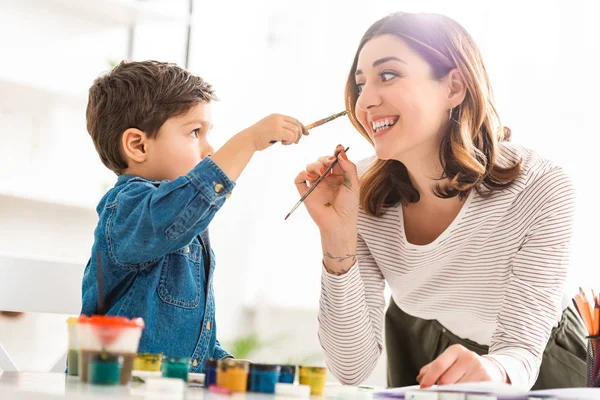 Felice madre e figlio divertirsi con pennelli vicino a contenitori con vernici — Foto stock
