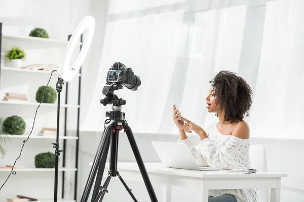 Attractive african american influencer holding smartphone near laptop and digital camera — Stock Photo