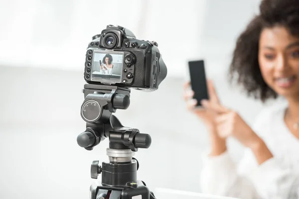 Mise au point sélective de l'appareil photo numérique avec un blogueur vidéo afro-américain attrayant dans des accolades pointant du doigt le smartphone avec écran blanc à l'écran — Photo de stock