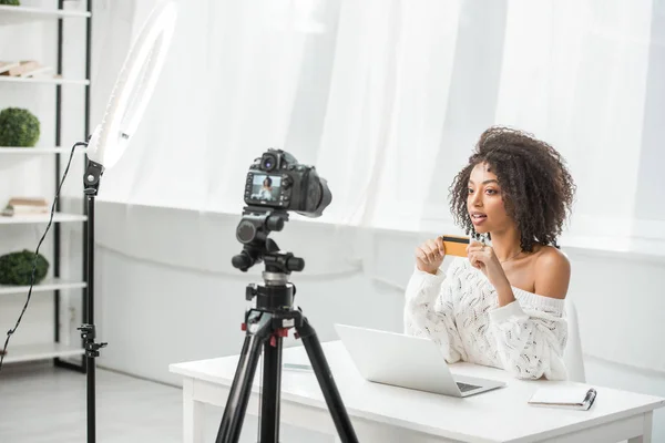 Foco seletivo de influenciador afro-americano atraente segurando cartão de crédito perto do laptop e olhando para a câmera digital — Fotografia de Stock